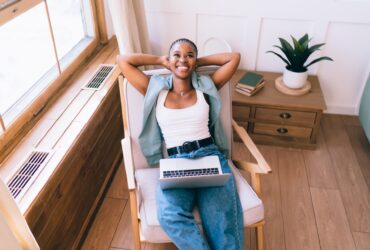 From above of thoughtful black female chilling and stretching arms up while sitting in armchair with wireless computer at home